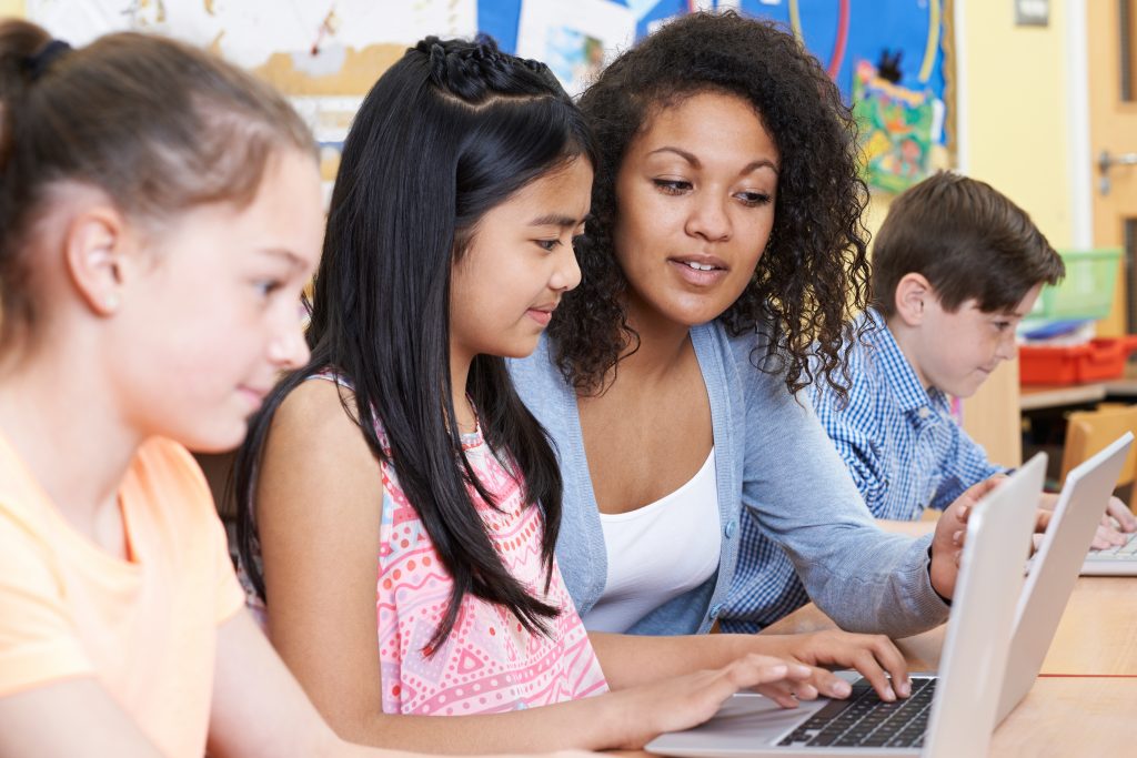 Teacher Helping Group Of Elementary School Children In Computer Class